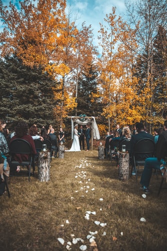 Aisle view of a couple saying "I do" at a fall wedding in front of trees covered in golden leaves.