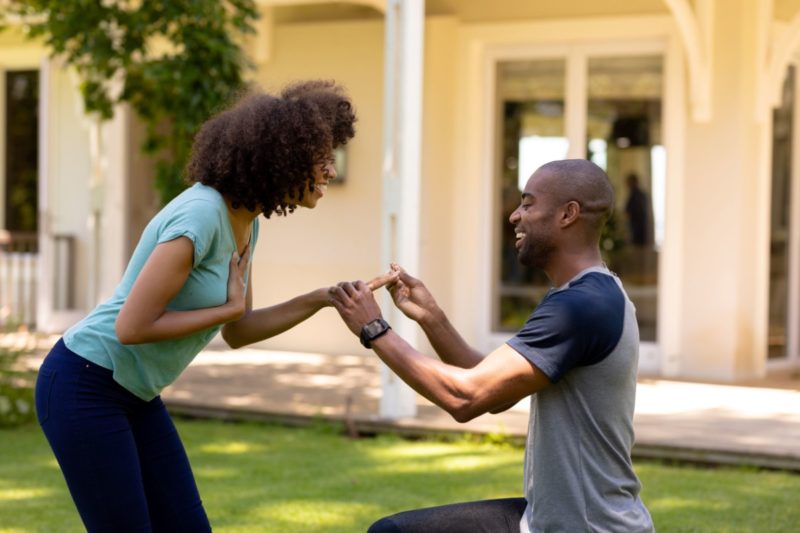 Traditions and customs - engagement proposal - man down on one knee in the garden as he places an engagement ring on her finger