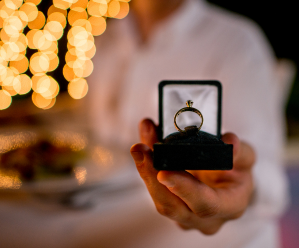 Engagement ring encased in a black velvet box being held by a man dressed in all white. In the foreground, blurry lights frame the moment showcasing the importance connected with the perfect proposal.