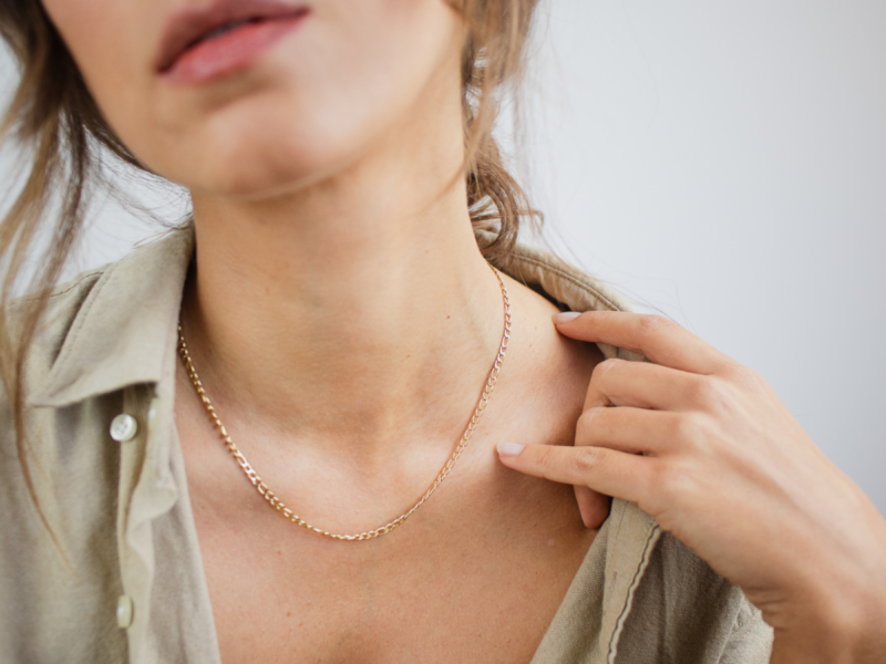 A woman with blond hair pulls her shirt collar back away from her neck showcasing her dainty gold chain necklace.